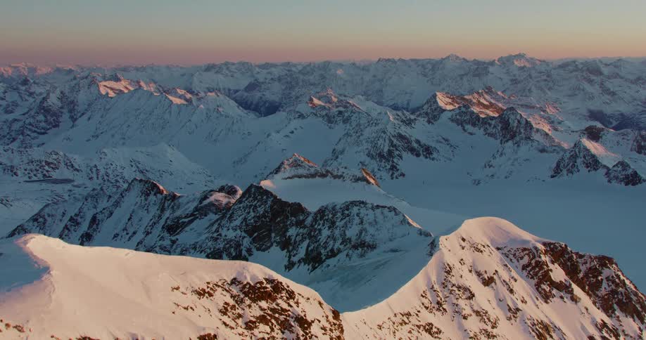 A lone cross is placed atop the peak of a mountain within a snow covered mountain range