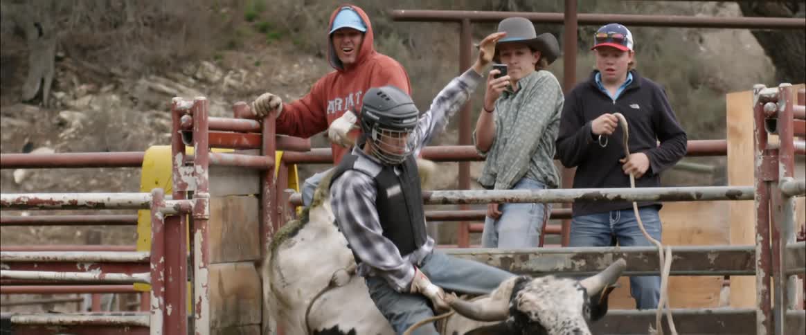 Group of people lean on a gate as a man opens it and a person riding a bull comes out