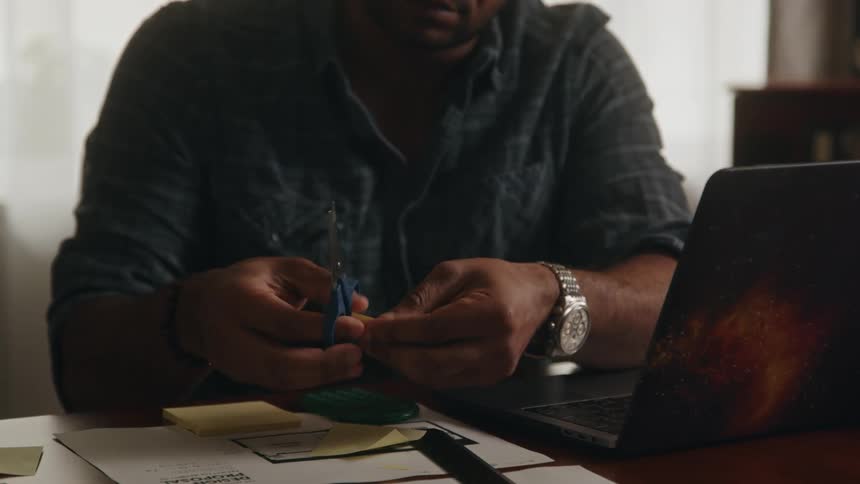 A man is sitting at a table with a laptop computer as he holds scissors and cuts paper into small pieces