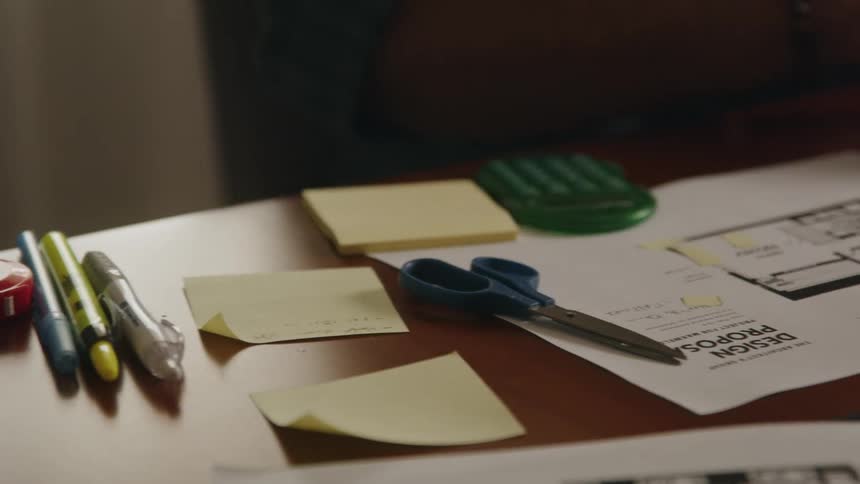 A man is sitting at a desk with a laptop computer and writing with a pencil on a note and there are scissors and pens on the table