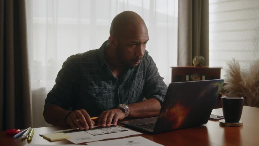 A man sits in his office and uses a ruler on paper as he looks at his laptop