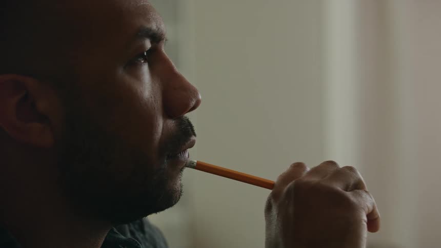 A man is sitting down thinking as he holds a pencil and presses it against his face