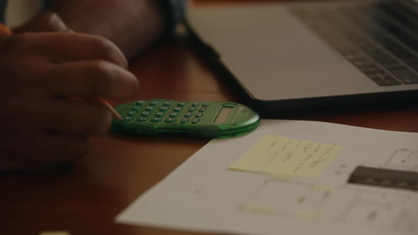 An up close of a persons hand holding a pencil typing on a calculator then writing on paper in front of them