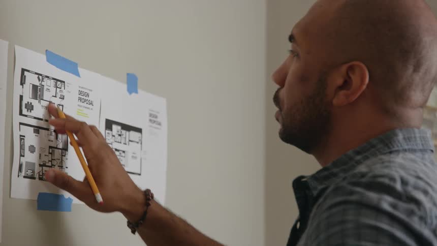 A man looks at design proposal papers taped to a wall and writes on them with a pencil