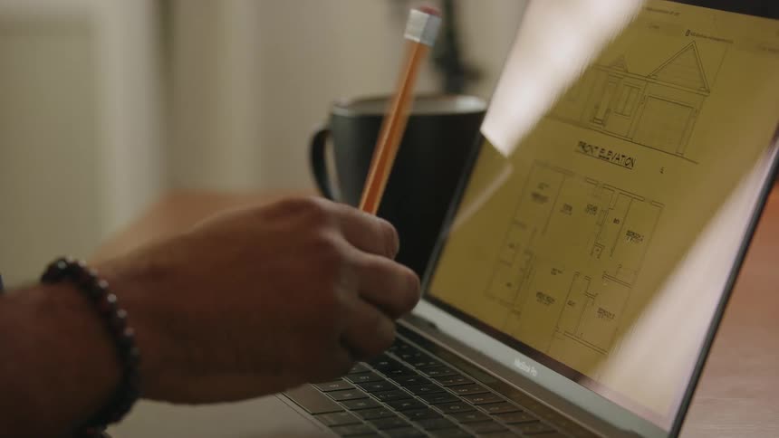 Male architect sitting at desk and working on first floor house plans on laptop