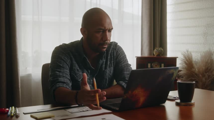 A man sits at a desk in his office and twirls a pencil in his fingers as he looks at his computer