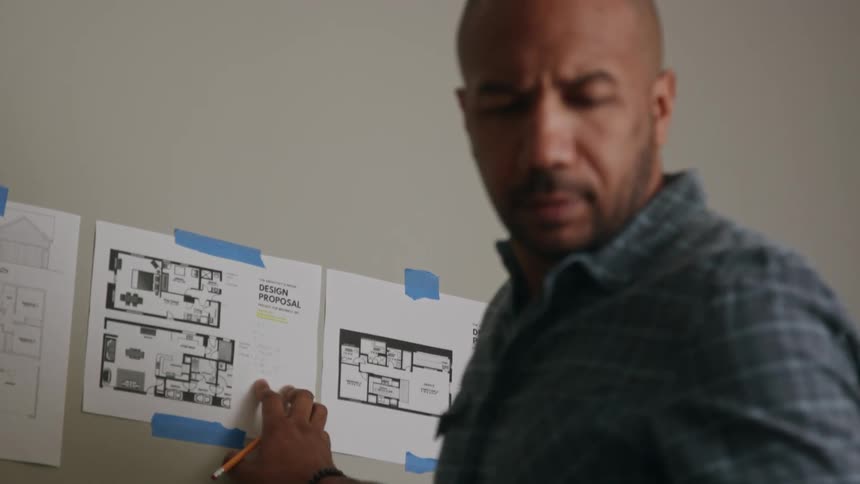 A man frowning looking down then turning to look at papers on the wall behind him