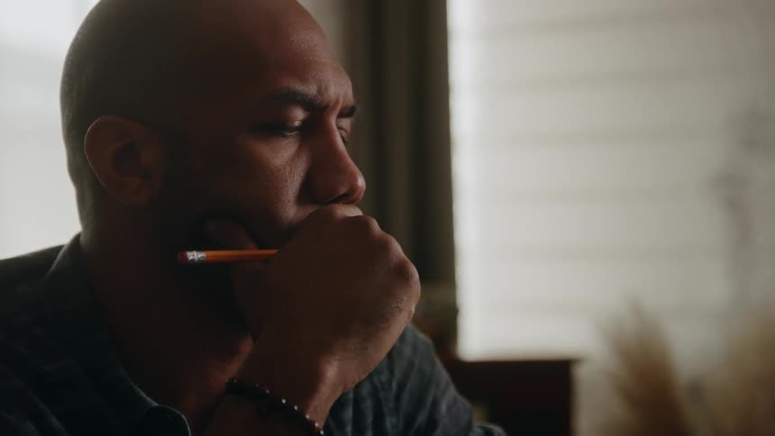A man sitting at a desk thinks about something and then writes with a pencil