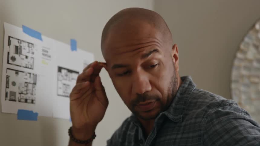 A man is writing on a chart taped to the wall and he sticks his pencil behind his ear