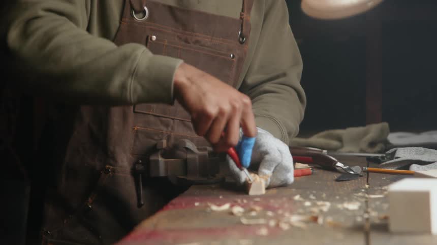 A man standing at a work bench using a tool to carve out a small piece of wood