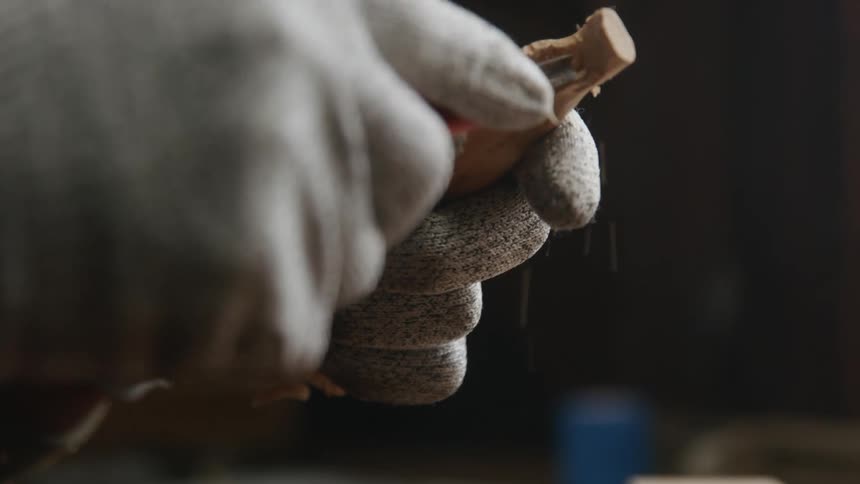 A person wearing gloves uses a blade to carve a piece of wood