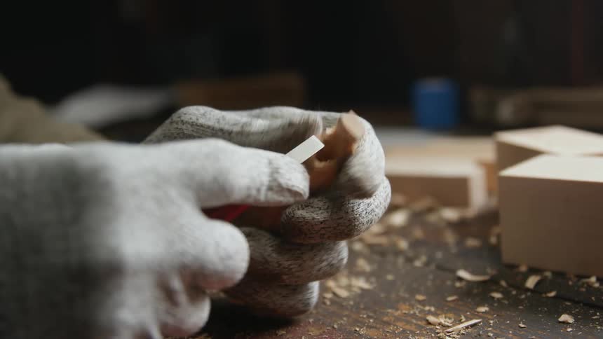 A man is wearing gloves as he uses a small chisel to shape a piece of wood