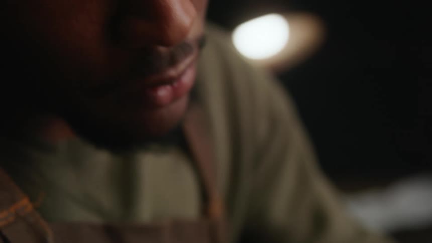 Woodcarver wears gloves while carving piece of wood in workshop lit by lamp