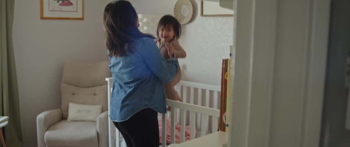 Pregnant mother with blue shirt and black pants takes toddler girl out of crib in room