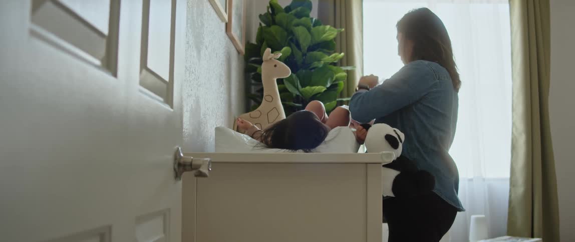 A pregnant mother changing her daughter on a changing pad on a table while the girl plays with a stuffed animal