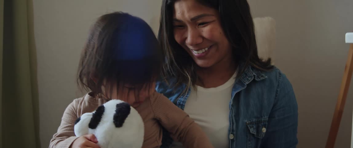 A little girl holding her stuffed panda is sitting with her mother as they smile and wave to the camera