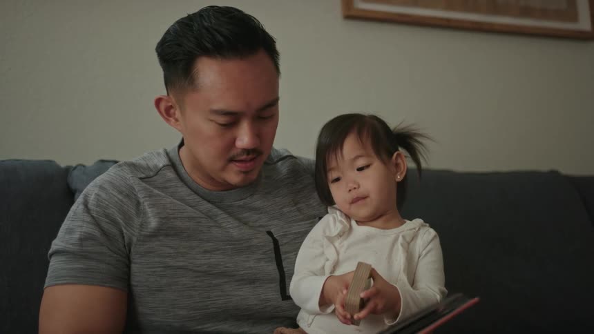 A father and daughter sit together on a couch looking at books
