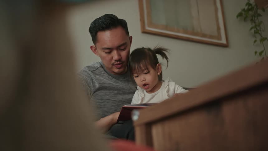 A man sits at home with his daughter and they read a book together