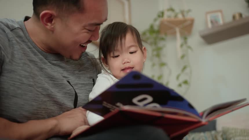 Father holds his daughter in his lap while reading to her taking a brief break to tickle her