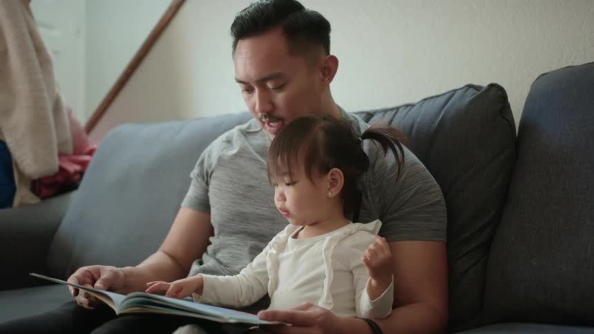 Father and daughter sit on couch while he reads a storybook to her