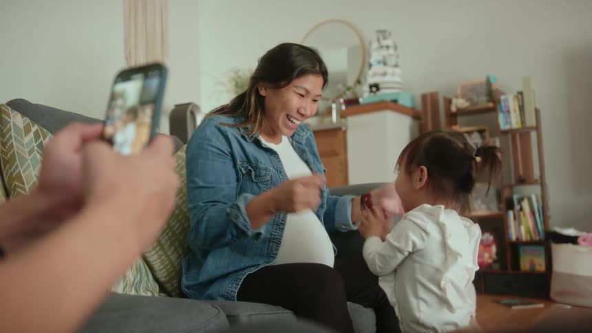 Father uses a smartphone to film his pregnant wife and baby daughter playing in the living room at home