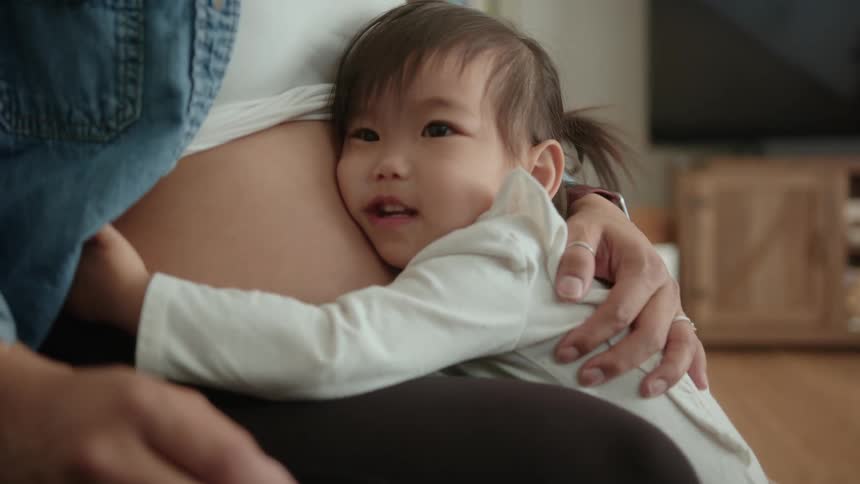 A little girl hugging her mothers pregnant belly while the mother sits