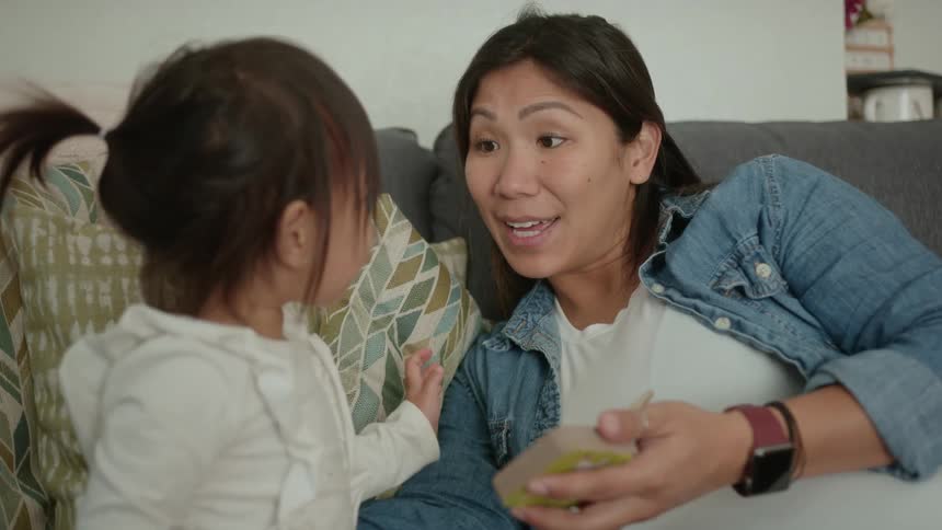 A mother sits on a couch with her daughter and holds a book as her daughter points at it
