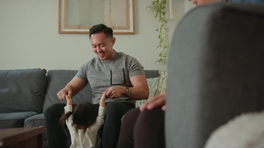 A man is sitting on the living room couch holding his young daughters hands as he helps her stand up and jump