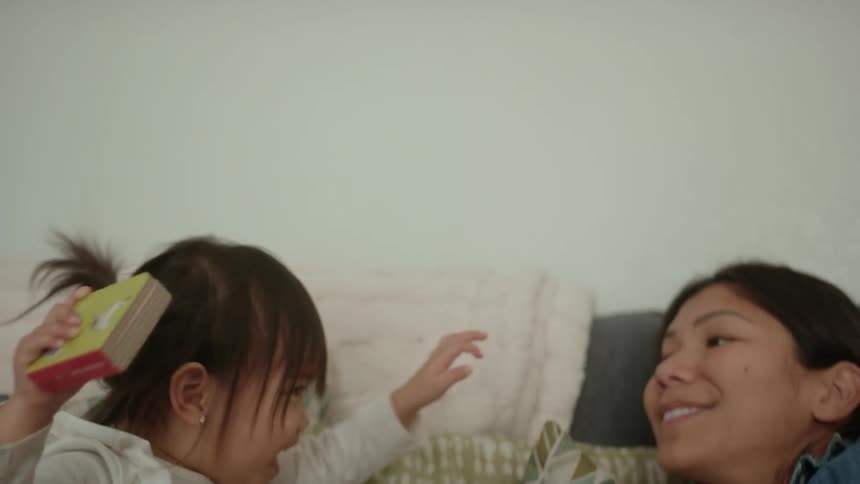 Daughter with white shirt and brown pants sits down next to pregnant mother lying on couch