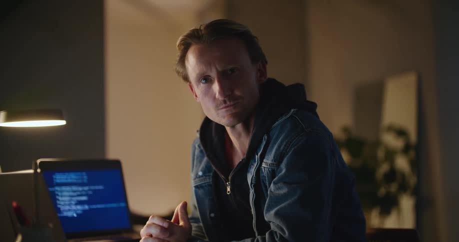 A man sitting at a desk in front of two laptop screens looking at the camera while thinking