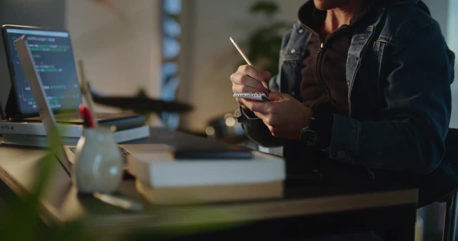 A man sitting at the office desk is thinking and then writing something on a piece of paper while looking at a laptop