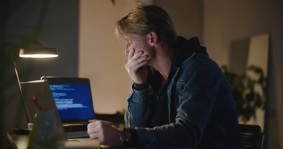 A man sits at his desk in his office and puts his head down after looking at his computer and rubs his eyes in frustration before typing on his laptop again