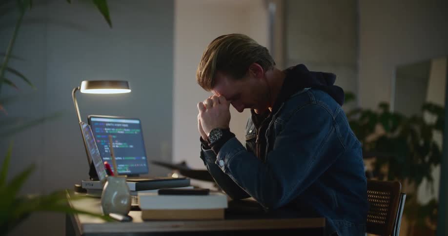 A man rubs his eyes while working on his laptop