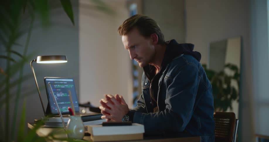 A man is sitting at a desk typing on a laptop computer and looking over at another screen as he types