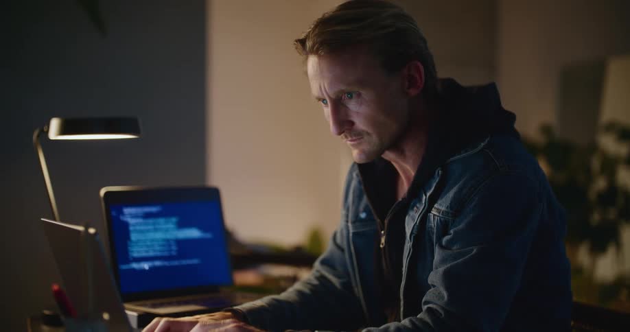 A man types on a laptop as lines of code process on a screen behind him