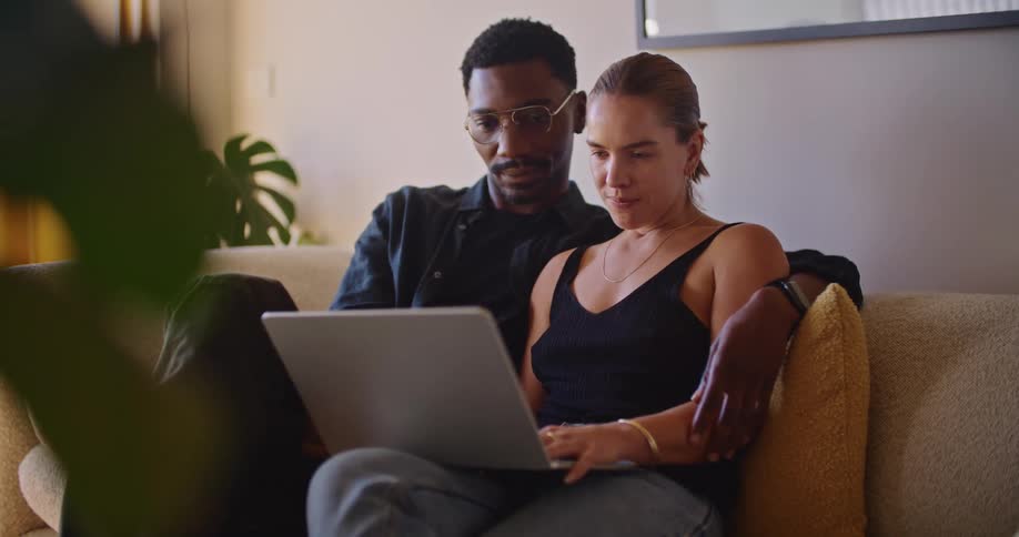 Man keeps arm around woman as they sit on couch talking and sharing a laptop