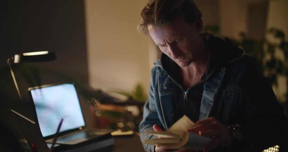 A man is sitting at a desk typing on a laptop computer and stops to look at a book before he continues to type