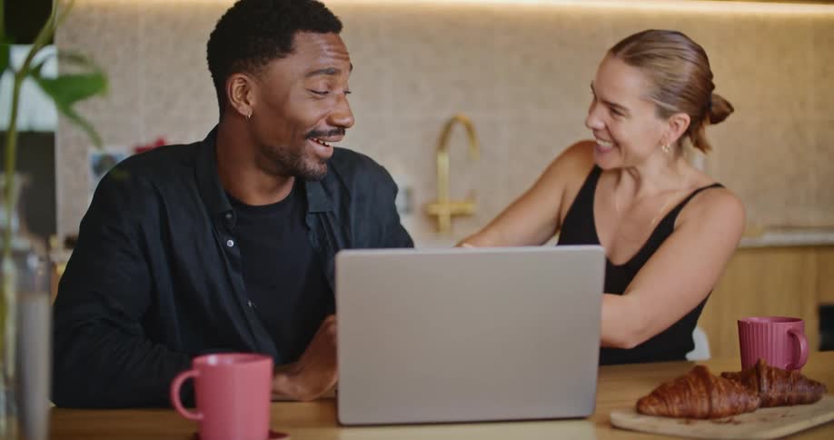 A couple are watching a laptop screen at the kitchen table and celebrating laughing and hugging each other