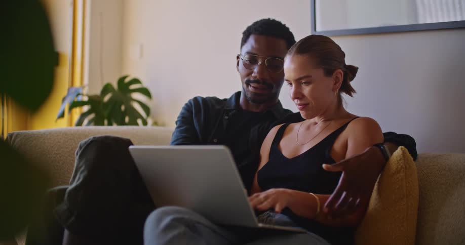 A romantic couple sitting on the couch have a conversation while doing work on a laptop