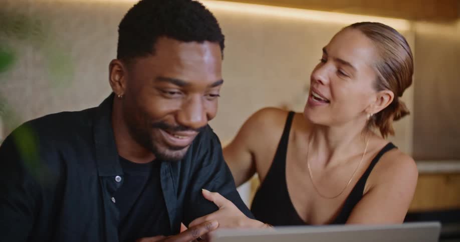A woman kisses her boyfriend on the cheek and hugs him while sitting at the dining room table and watching the screen of a laptop computer as they celebrate good news