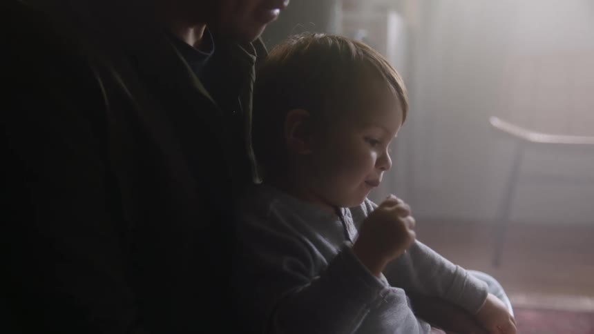 A father holds his son on his lap while the boy eats something and smiles shaking his head