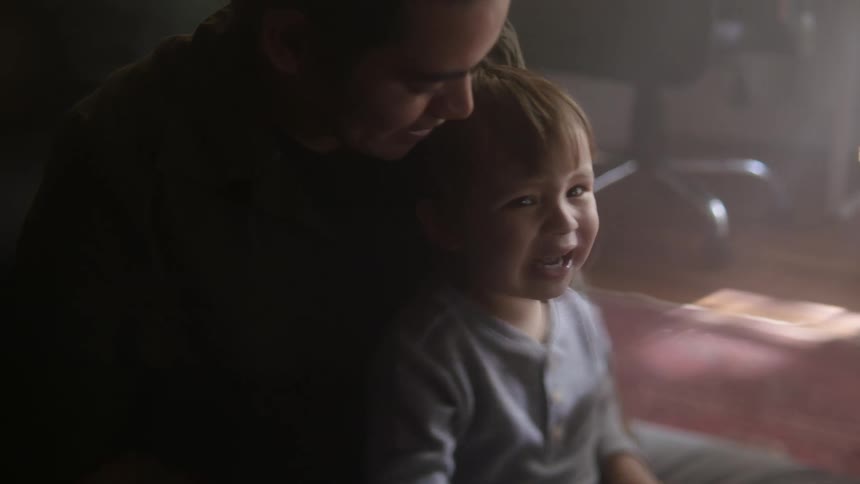 Toddler boy sits on fathers lap laughing and talking to mother