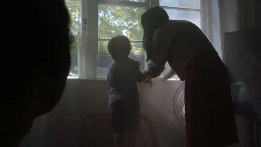 A mother and young son stand in front of a window looking out while a grandmother sits back and watches