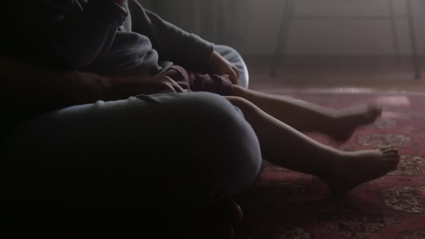 A baby is sitting on hisher moms lap on the floor in the morning kicking its little heels against a red carpet on the floor