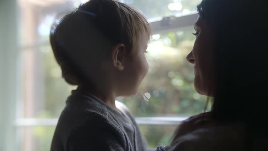 Young son reacts in surprise as his mom holds him while standing near window talking with him