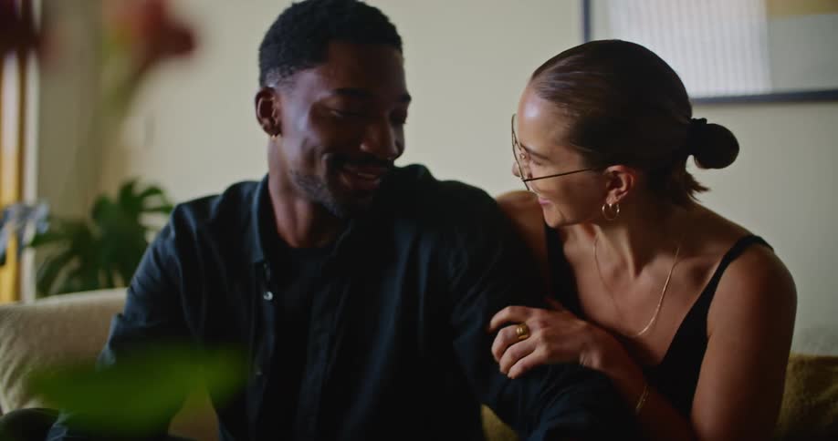 A man and a woman are sitting on the couch together talking and smiling and she pats him on the back