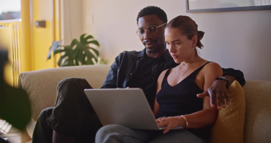 A young couple are sitting on the sofa in the living room chatting and watching a laptop
