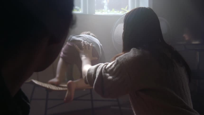 A mother helps her son get up on a chair in front of a window next to a fan