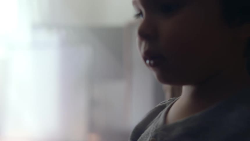 A little boy is walking across the room as his mother sits in the distance