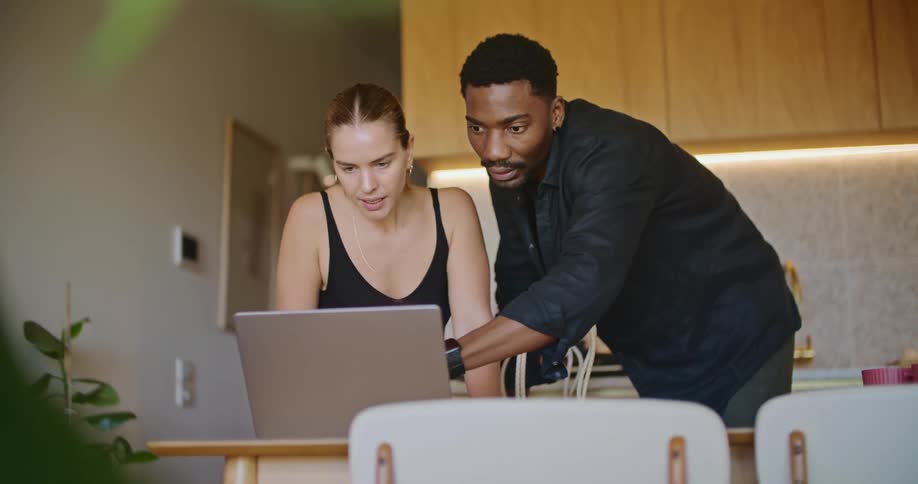 A man and a woman are standing over the kitchen table looking at a laptop computer and pointing at the screen as they talk
